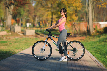 Obraz premium Young beautiful girl in sportswear stands next to a bicycle on the street in the park in autumn.