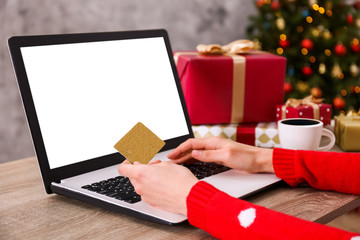Female hands with golden credit card, laptop, christmas presents, decorated tree on background. Woman picking presents online, eshopping on her laptop. Wooden table, copy space, close up.