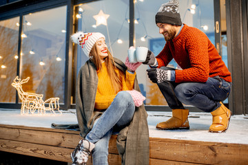 Young couple warming up with plaid and hot drinks sitting on the terrace of the modern house in the mountains durnig the winter holidays