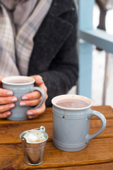 Two cups of cocoa on a wooden table, one of which is held in the hands of a woman in a warm coat. Drink a hot drink on the open terrace in winter. Nature and relaxation on a snowy day