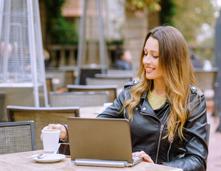 Happy young woman with hot drink using laptop in outdoor cafe