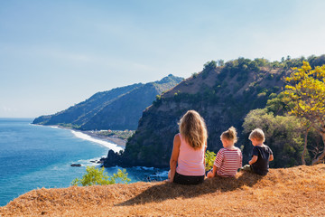 Fototapeta na wymiar Family vacation lifestyle. Happy mother, kids on hill with scenic view of high cliffs, fishers village on black beach. Children looking at blue sea. Bukit Asah is popular travel destination in Bali.