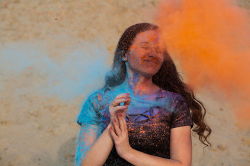 Cheerful brunette woman with long curly hair playing with exploding orange and blue dry Holi powder