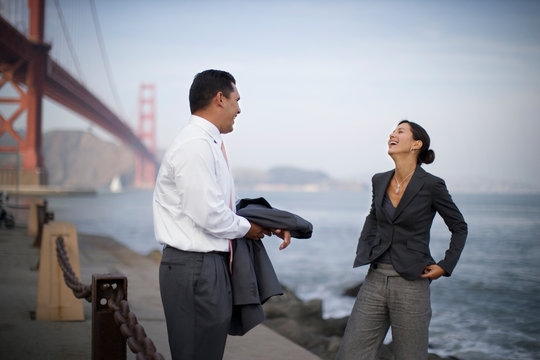 Laughing businessman and businesswoman having a conversation while standing at the edge of a waterfront overlooking a harbor.