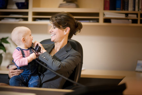 Smiling mid-adult businesswoman holding a telephone receiver to her young baby's ear while seated at a desk in her office.