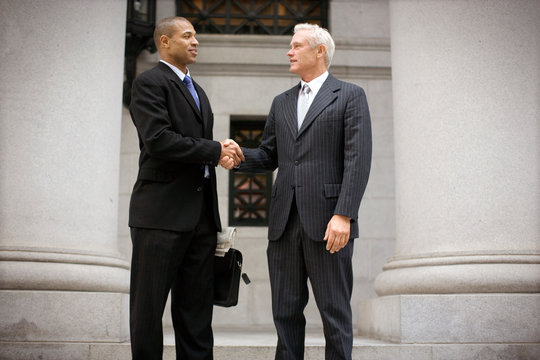 Two suited businessmen shaking hands outside a building in the city.