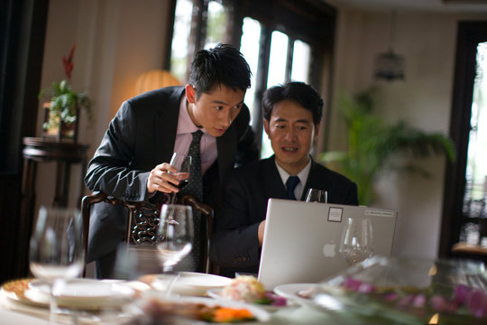 Two businessmen using a laptop while drinking wine at a table inside a restaurant.