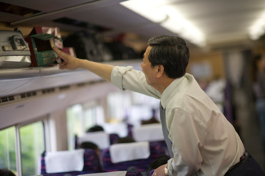 Mature adult businessman packing a bag in an overhead locker on a train.