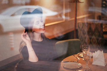 Beautiful young smiling and happy woman blonde in a hat at a restaurant table through the glass. Reflections