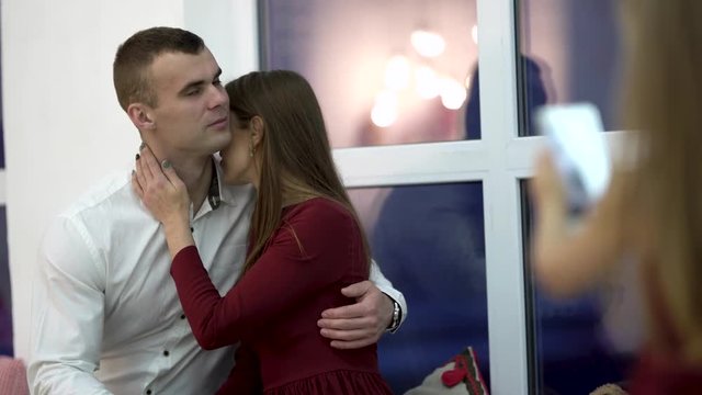 Pretty small blond girl taking photo of her young loving parents on Christmas. Wife kissing husband on cheeck and tightly hugging him. New Year love atmosphere. Big window background
