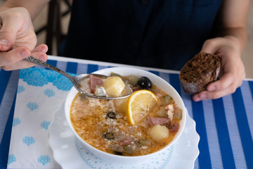 Traditional Russian soup Solyanka cooked with meat, sausages, salted cucumbers on wite bowl. Black bread, lemon, olived and tomatoes on color blue background, homemade food.