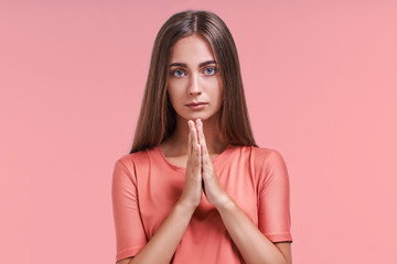 Fototapeta premium Horizontal portrait of pretty woman keeps palm together, has pleased expression, asks husband to buy her new coat. Glad attractive female makes request, pleads for mercy. Hopeful young adult.