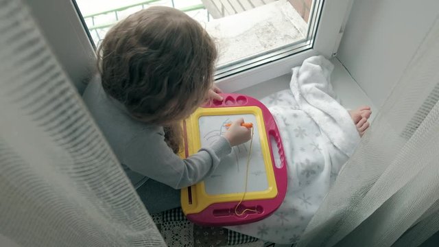 A little girl with red wavy hair sits on the windowsill and draws on a magnetic board. The concept of the educational process.