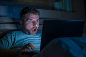 attractive internet addict man in shock and surprise networking late night on bed surprised face expression working with laptop computer in social media addiction - obrazy, fototapety, plakaty