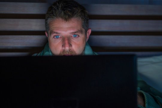 attractive and relaxed internet addict man networking concentrated late at night on bed with laptop computer in social media addiction or workaholic businessman concept