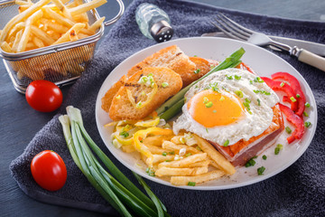 Fried egg with bacon in a white plate with fried pieces of bread, greens, tomatoes and french fries on a gray wooden table. Close-up