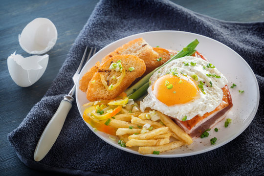 Fried egg with bacon in a plate with fried pieces of bread, greens and french fries on a gray wooden table. Close-up