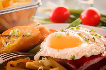 Fried egg with bacon in a white plate with fried pieces of bread, greens, tomatoes and french fries on a gray wooden table. Close-up