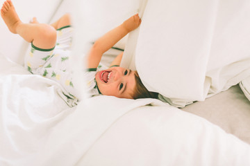 A little boy laughing in the sheets of a white bed. 