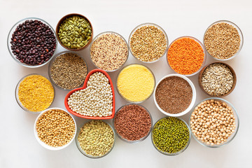 Top view to glass bowls with cereals, beans and seeds with red heart-shaped bowl in the middle on white background.