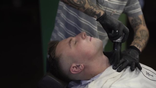 Tattoed  barberman in black gloves applying some powder using big brush on the face of male customer and spraying some liquid on his face. Man's covered with white peignoir