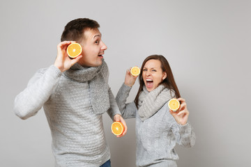 Fun couple girl guy in gray sweaters, scarves together hold orange lemon isolated on grey wall...