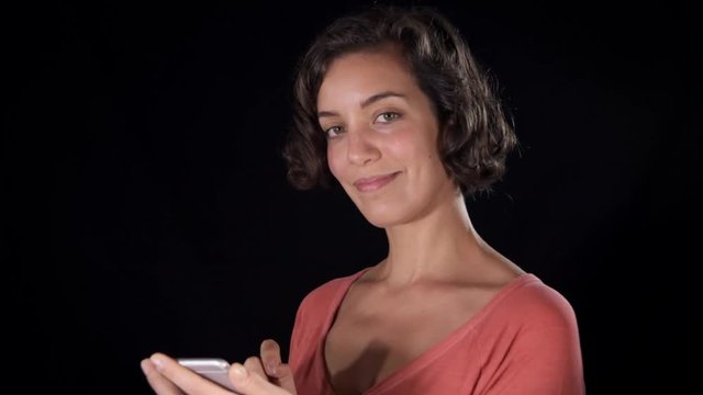 Young woman, isolated on a black studio backdrop, plays with her phone and after some hesitation, presses the screen decisively