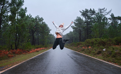 Una chica joven y deportista salta en un lluvioso día de invierno en el medio de una carretera dentro de un bosque frondoso