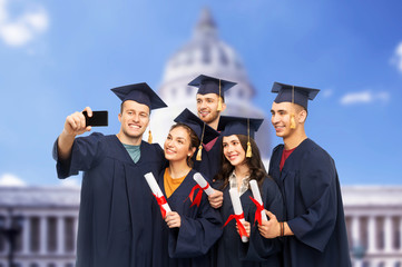 education, graduation and people concept - group of happy graduate students in mortar boards and bachelor gowns with diplomas taking selfie by smartphone over washington white house background
