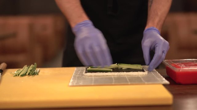 Close-up shot of man's hands putting cucumber on rice to make sushi roll.