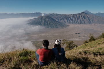 Fototapeta na wymiar Back view of Asian couple sitting on the hill and looking to the sea of fog over Bromo Tengger Semeru National Park in East Java, Indonesia. Early morning