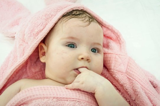 Sweet Caucasian newborn 3 months old baby girl wrapped in a pink bath towel with a finger in her mouth looking at the photographer camera.