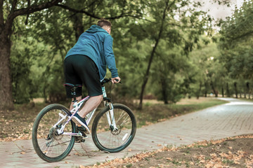 Fototapeta premium Young caucasian man bicycles through the abandoned path in the autumn park, casual sportswear. Dark hair, white earphones. Outdoors, copy space. Favorite music or audio book, morning fitness