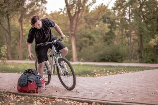 Attractive young caucasian man with dark hair on the bicycle in the park, leans towards his bag with sportswear. Outdoors, lovely autumn (fall) day. Copy space