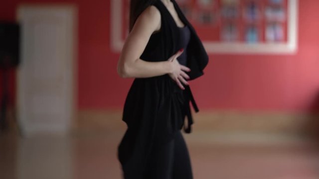 Close-up shot of woman's upper body part. Slim and  sexy dancerl in black dress is performing Latin American dance in ballroom. Back view of red wall with photos.
