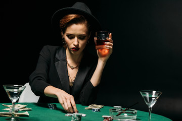 beautiful girl in jacket and hat holding glass of whiskey and playing poker in casino