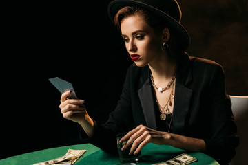 attractive girl in jacket and hat looking at poker cards and holding glass of whiskey in casino