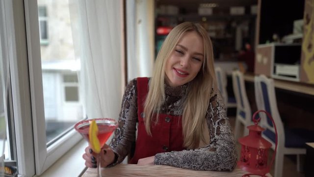 Young smiling woman is sitting in cafe with a red-coloured cocktail, looking at the camera and talking. Concept of the friendly, cozy chat. Smiling woman talking to the camera as with her friend.