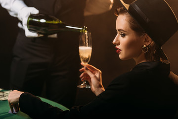 side view of waiter pouring champagne in glass of beautiful girl in jacket and hat in casino
