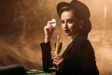 attractive girl in jacket and hat holding glass of champagne and poker chips at table in casino