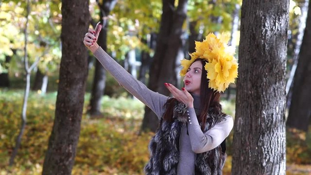 The girl picks herself. Makes a selfi. In the autumn park with a wreath of yellow leaves on the head