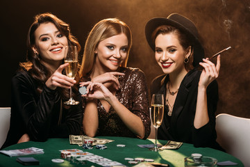attractive girls with glass of champagne, cigarette and poker chips sitting at table in casino and...