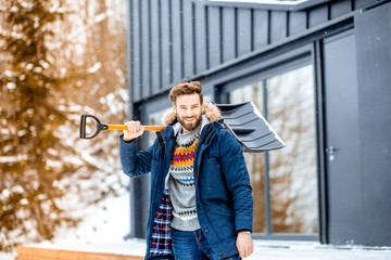 Portrait of a man in winter clothes with a snow shovel near the modern house in the mountains