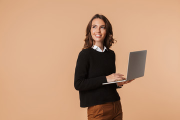Photo of cute woman 20s standing and holding laptop, isolated over beige background