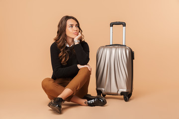 Photo of displeased woman sitting on floor with baggage, while isolated over beige background