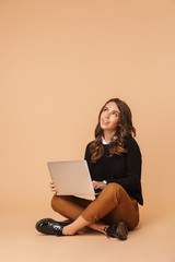 Image of charming woman 20s using laptop, while sitting on floor isolated over beige background