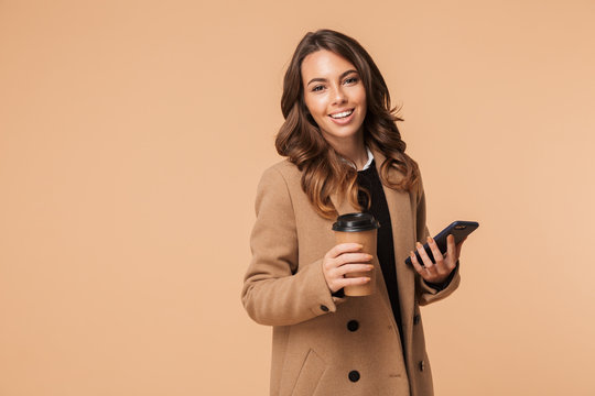 Image of pretty woman 20s wearing coat holding mobile phone and takeaway coffee while standing, isolated over beige background