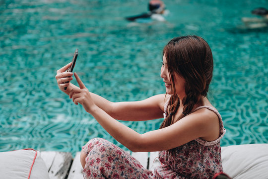 Vacation and technology. Colorful portrait of pretty young woman taking selfie portrait with her smartphone near swimming pool on tropical beach. 