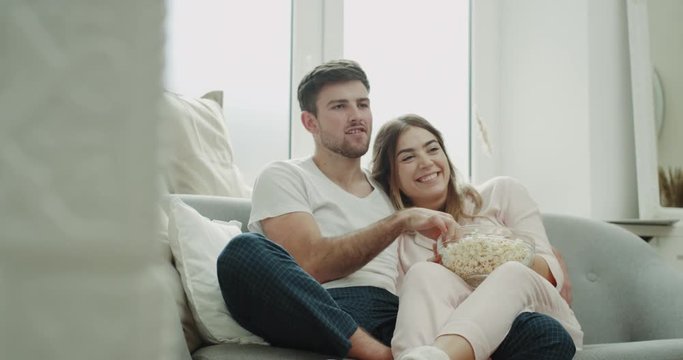 Romantic time hugging couple in pajamas sitting on the sofa and watching tv eating popcorn and happy spending morning together.