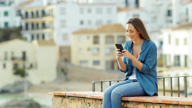 Happy woman uses smart phone and her partner arrives and share it sitting on a ledge on vacation